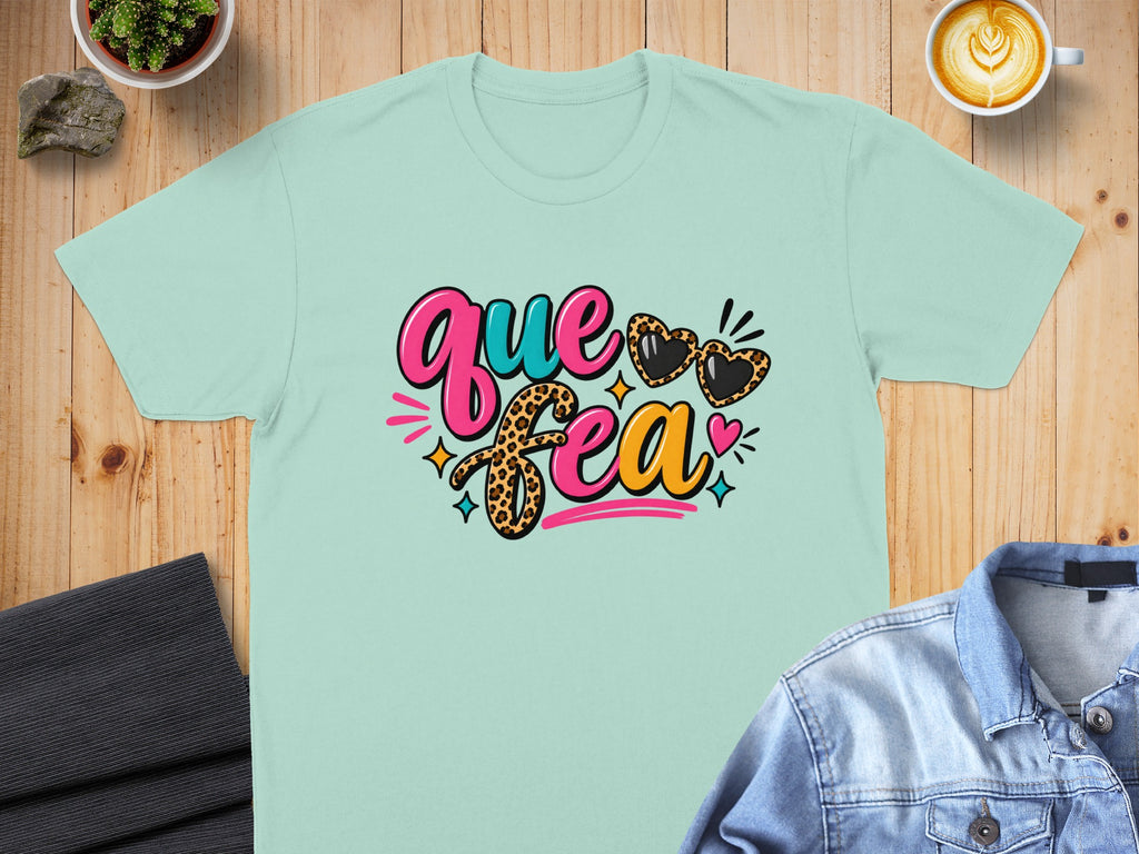 Light teal t-shirt with colorful "que sera" graphic and heart-shaped sunglasses.
