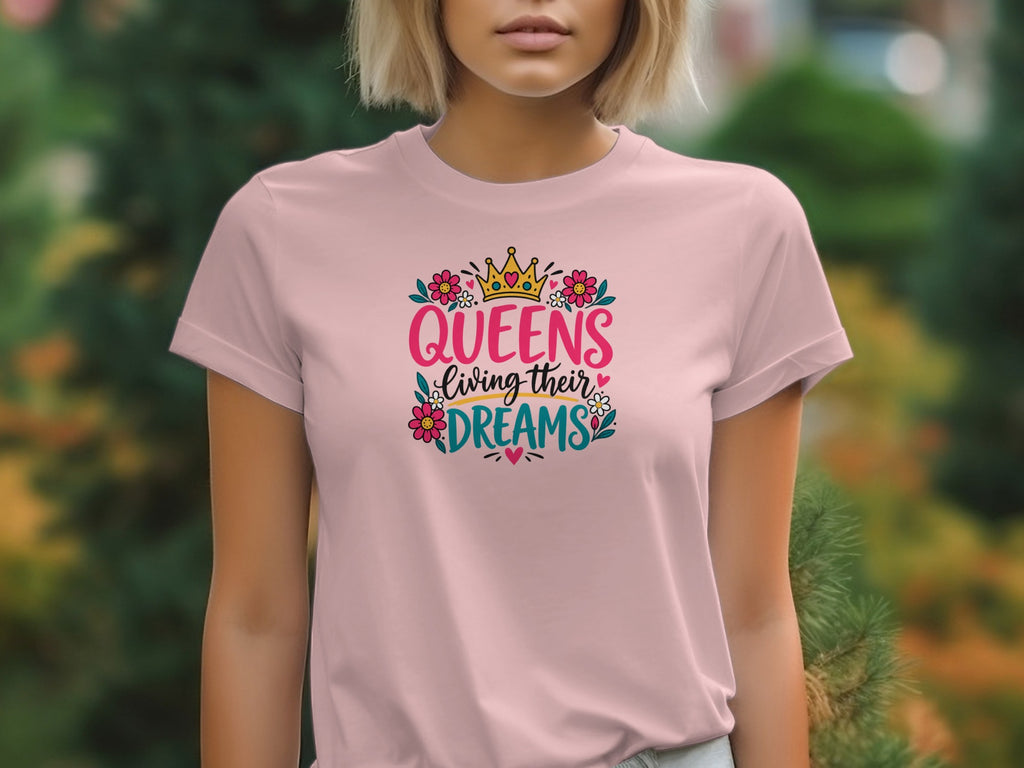 A person wearing a pink t-shirt with the graphic text QUEENS living their DREAMS, crowned with flowers and hearts.